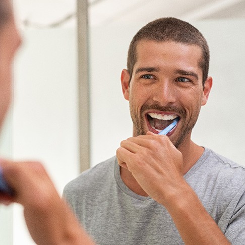 A smiling man brushing his teeth in front of a bathroom mirror